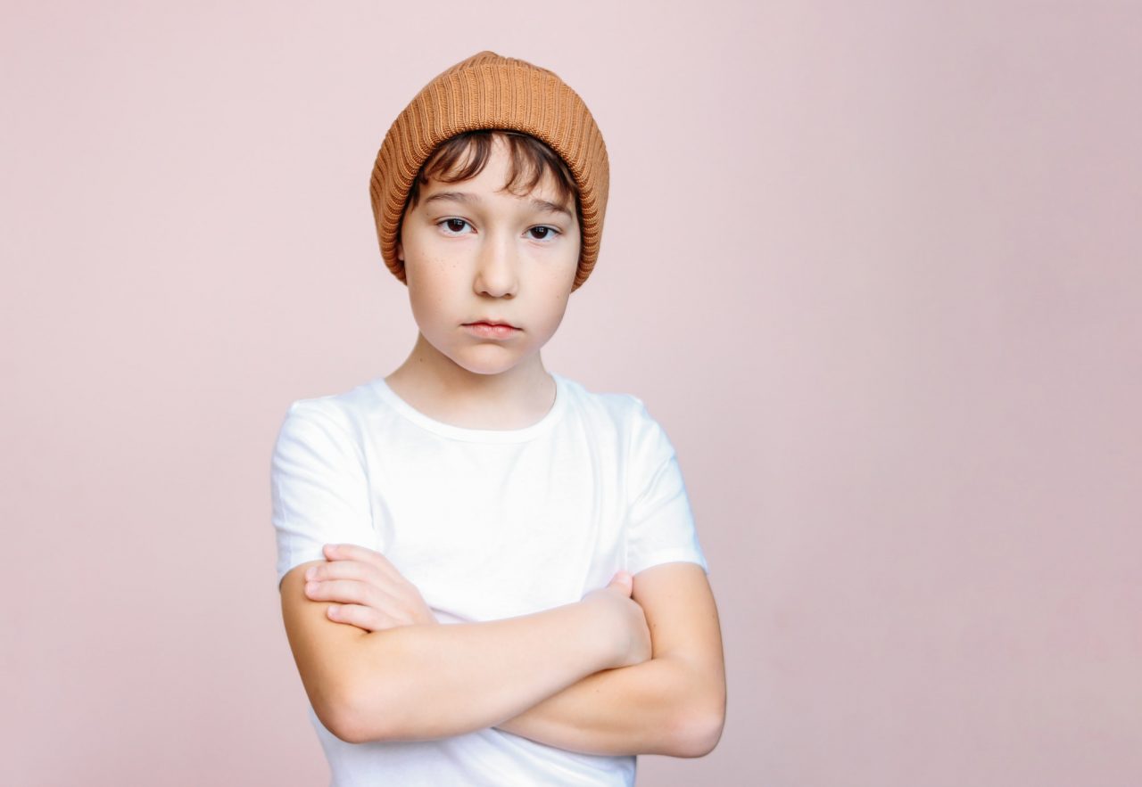 attractive tween boy with dark hair in hat looks at camera on light pink background